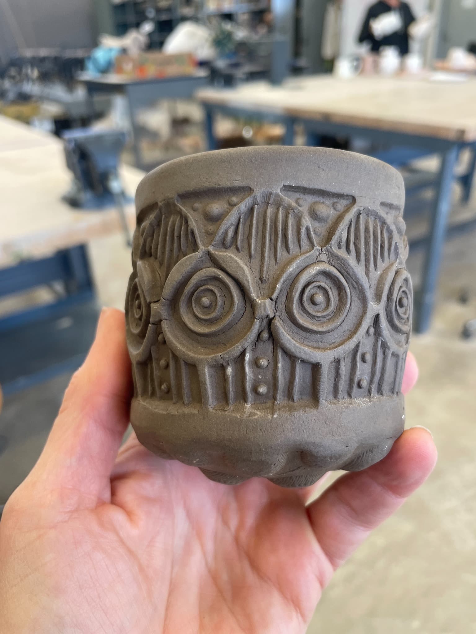 A hand holding a raw dark clay cup with deeply stamped circular and geometric patterns.